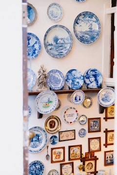 A collection of decorative blue and white ceramic plates is displayed on a wall. The plates feature various designs, including intricate patterns, landscapes, and portraits. Additional items such as framed pictures, a small statue, and other decorative objects complement the plate arrangement.