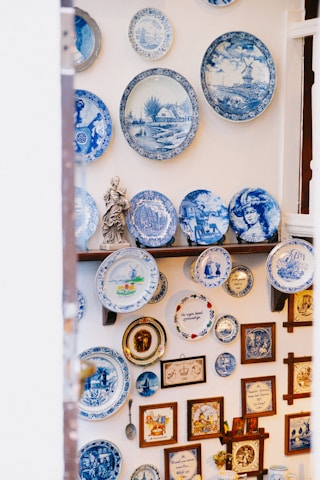 A collection of decorative blue and white ceramic plates is displayed on a wall. The plates feature various designs, including intricate patterns, landscapes, and portraits. Additional items such as framed pictures, a small statue, and other decorative objects complement the plate arrangement.