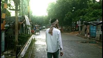 A person wearing a white shirt and a headscarf walks down a narrow, rural road lined with makeshift shelters and greenery. The sky is overcast, creating a subdued and tranquil atmosphere. An auto-rickshaw and other vehicles are visible in the distance, suggesting a lightly trafficked area.