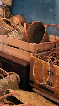 A collection of woven baskets with handles are neatly arranged in wooden and cardboard boxes. The baskets vary in shape and size, showcasing intricate weaving patterns. The earthy tone of the baskets contrasts with the muted background wall.