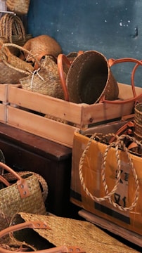 A collection of woven baskets with handles are neatly arranged in wooden and cardboard boxes. The baskets vary in shape and size, showcasing intricate weaving patterns. The earthy tone of the baskets contrasts with the muted background wall.