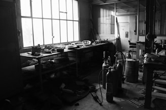 A dimly lit industrial workspace with large windows allowing some natural light. The room contains cluttered shelves with tools, equipment, and various materials. Fire extinguishers and other safety equipment are visible, and there is a general sense of disarray.