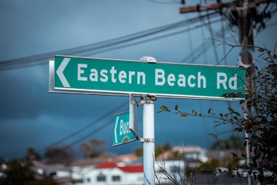 A road sign indicating 'Eastern Beach Rd', set against a backdrop of power lines and an overcast sky. Sparse branches and leaves are visible in the foreground, adding a touch of nature to the urban setting.