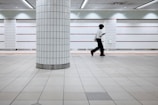 A person is walking in a tiled underground passageway. The floor and walls are made of white tiled panels with orange and black accents. A single large column stands in the foreground. The person appears to be focused on a phone or device, adding a sense of modern urban lifestyle.