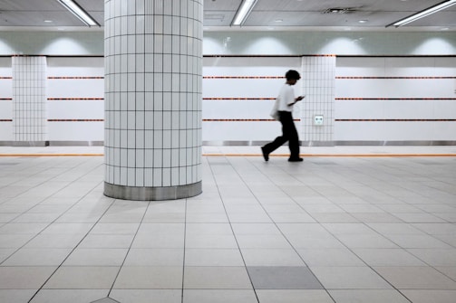 A person is walking in a tiled underground passageway. The floor and walls are made of white tiled panels with orange and black accents. A single large column stands in the foreground. The person appears to be focused on a phone or device, adding a sense of modern urban lifestyle.