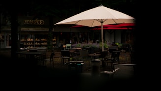 An outdoor seating area with tables and chairs under large umbrellas, set in a dimly lit environment. There are plants and a shop in the background, suggesting a quiet and cozy atmosphere.