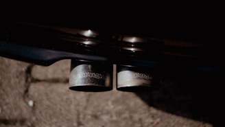 A close-up view of a car’s dual exhaust pipes with visible branding on them. The pipes are metallic and polished, set against a dark background with some pavement visible.