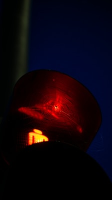 A close-up view of a red traffic light with a web spanning across its surface. The background appears dark, suggesting it is nighttime or low light conditions.