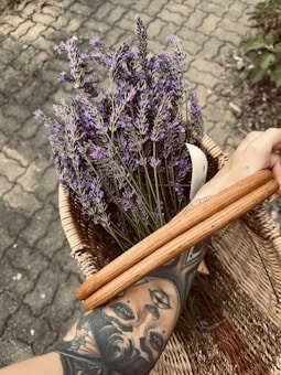 A tattooed arm holding a basket filled with fresh lavender sprigs. The basket features a wooden handle and rests on a stone pavement with a brick pattern.