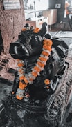 A black stone sculpture of a bull adorned with an orange and white marigold garland sits on a pedestal. The setting appears to be indoors, with stone walls and a donation box in the background.