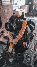 A black stone sculpture of a bull adorned with an orange and white marigold garland sits on a pedestal. The setting appears to be indoors, with stone walls and a donation box in the background.