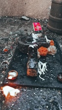 An outdoor ritual setup on a rough surface with a dark stone object, surrounded by white flowers and bright orange marigolds. A small lit oil lamp sits nearby, and a splash of white liquid is visible. In the background, a red package and some debris are scattered.