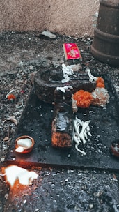 An outdoor ritual setup on a rough surface with a dark stone object, surrounded by white flowers and bright orange marigolds. A small lit oil lamp sits nearby, and a splash of white liquid is visible. In the background, a red package and some debris are scattered.