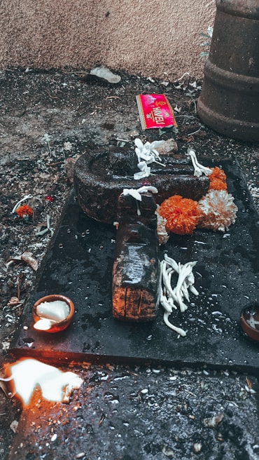 An outdoor ritual setup on a rough surface with a dark stone object, surrounded by white flowers and bright orange marigolds. A small lit oil lamp sits nearby, and a splash of white liquid is visible. In the background, a red package and some debris are scattered.