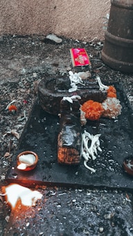 An outdoor ritual setup on a rough surface with a dark stone object, surrounded by white flowers and bright orange marigolds. A small lit oil lamp sits nearby, and a splash of white liquid is visible. In the background, a red package and some debris are scattered.