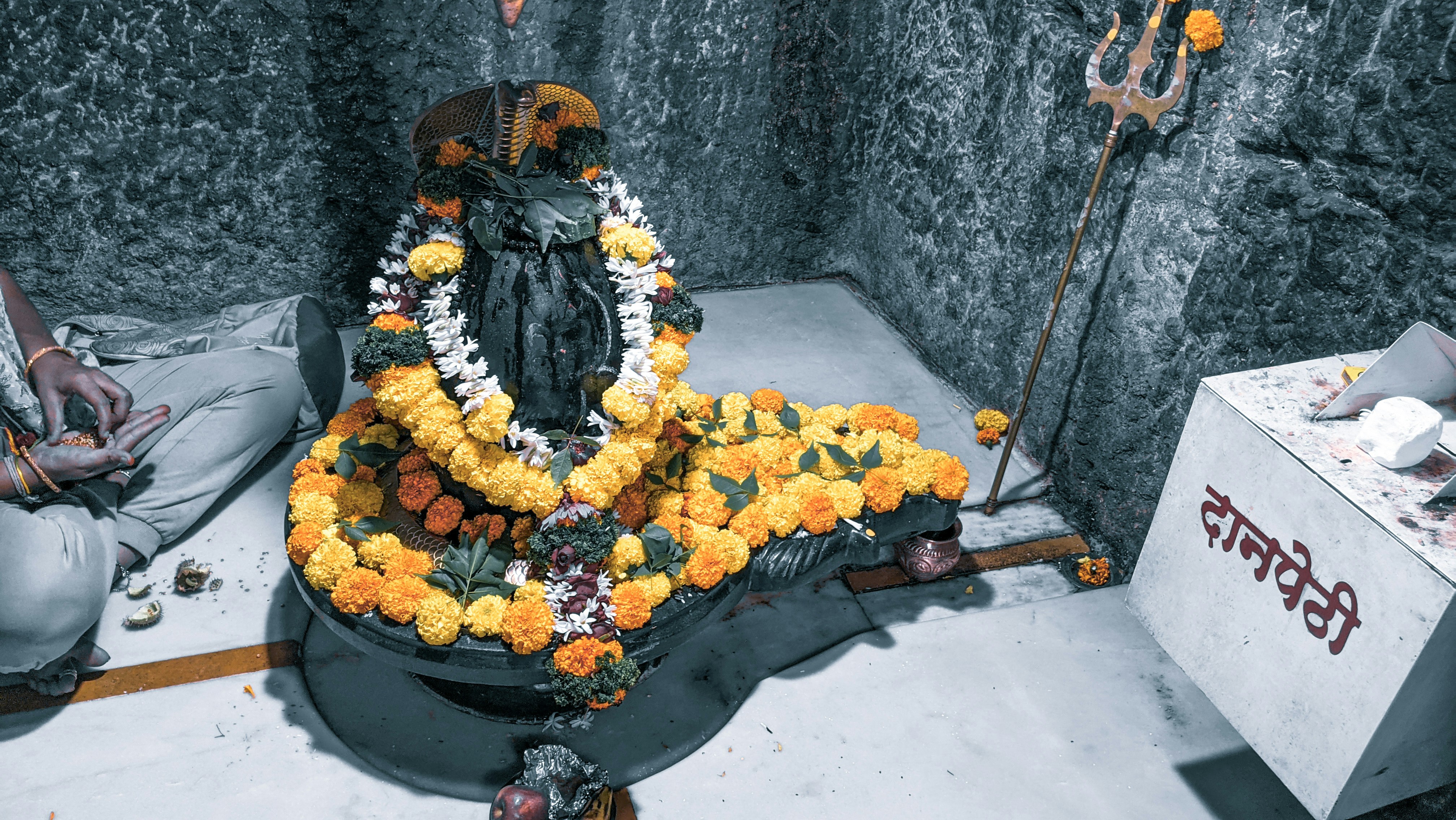 Shiva lingam adorned with marigold flowers and sacred offerings, soft amber candlelight, deeply shadowed temple sanctum, rich ochre stone walls