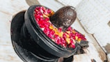 A stone lingam adorned with vibrant red, yellow, and pink flower petals is placed within a circular pedestal. The backdrop features tiled walls with a subtle pattern, and the marble floor adds a clean, sacred setting.