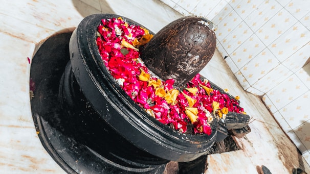 A stone lingam adorned with vibrant red, yellow, and pink flower petals is placed within a circular pedestal. The backdrop features tiled walls with a subtle pattern, and the marble floor adds a clean, sacred setting.