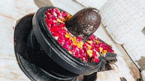 A stone lingam adorned with vibrant red, yellow, and pink flower petals is placed within a circular pedestal. The backdrop features tiled walls with a subtle pattern, and the marble floor adds a clean, sacred setting.