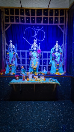 Three statues of deities adorned with garlands and traditional attire stand in front of a blue backdrop with the 'Om' symbol. The area is decorated with flowers and religious offerings placed on a table covered with a turquoise cloth.