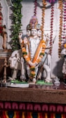 A statue of a four-faced deity adorned with colorful floral garlands and a decorative headdress. The deity holds a trident and various other items in its multiple hands and stands on a platform with a white cow beside it. The background features hanging strings of marigold and other flowers, along with a leafy vine on one side.