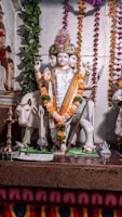 A statue of a four-faced deity adorned with colorful floral garlands and a decorative headdress. The deity holds a trident and various other items in its multiple hands and stands on a platform with a white cow beside it. The background features hanging strings of marigold and other flowers, along with a leafy vine on one side.
