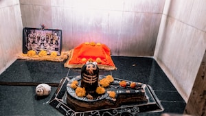 A sacred altar featuring a Shiva Lingam placed on a decorated base, adorned with yellow and orange marigold flowers. Behind it, there is a red cloth with additional marigolds, and a framed black-and-white photo of several deities or spiritual leaders. The altar is set against a backdrop of pale stone walls.