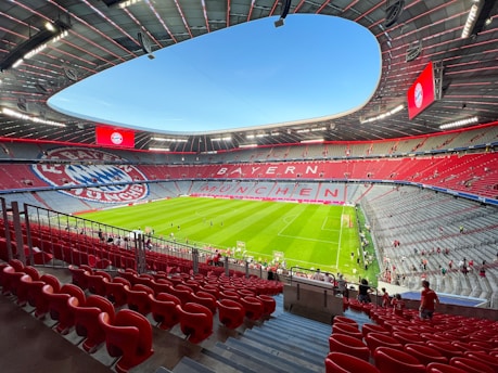 A large, modern football stadium with an expansive green field. The seating area is predominantly red, with sections of grey and the logo of a sports club visible. The words 'BAYERN MÜNCHEN' are prominently displayed in the stands. A few people are scattered around the seating area, suggesting an event is about to start or has recently ended.