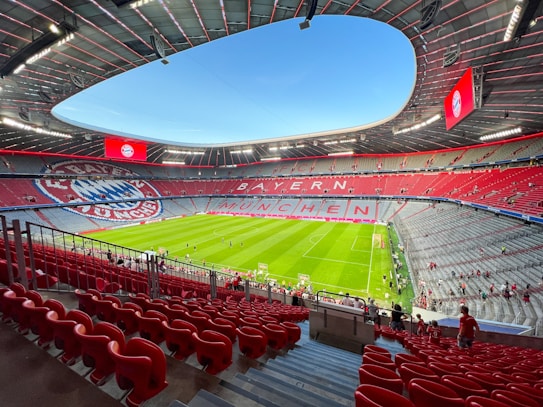 A large, modern football stadium with an expansive green field. The seating area is predominantly red, with sections of grey and the logo of a sports club visible. The words 'BAYERN M&Uuml;NCHEN' are prominently displayed in the stands. A few people are scattered around the seating area, suggesting an event is about to start or has recently ended.
