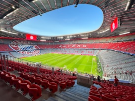 A large, modern football stadium with an expansive green field. The seating area is predominantly red, with sections of grey and the logo of a sports club visible. The words 'BAYERN MÜNCHEN' are prominently displayed in the stands. A few people are scattered around the seating area, suggesting an event is about to start or has recently ended.