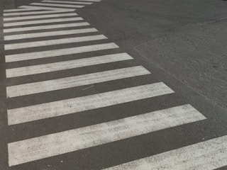 A crosswalk with white stripes is painted on a paved road surface. The paint appears slightly worn and the road is dark, possibly asphalt. The crosswalk forms a distinctive pattern against the background.