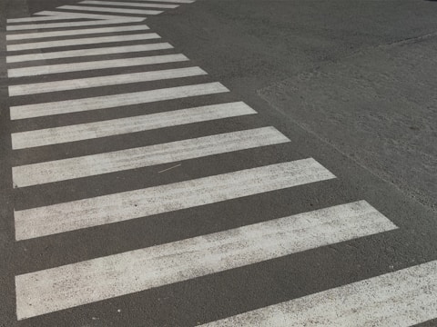 A crosswalk with white stripes is painted on a paved road surface. The paint appears slightly worn and the road is dark, possibly asphalt. The crosswalk forms a distinctive pattern against the background.
