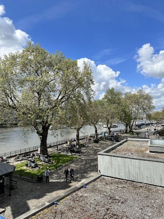 A scenic riverside view with large, leafy trees lining a path alongside a body of water. People are strolling along the walkway, which is bordered by a fence. There are benches and grassy areas under the trees, providing a space for relaxation. The sky is bright blue with fluffy white clouds, enhancing the peaceful and natural ambiance.