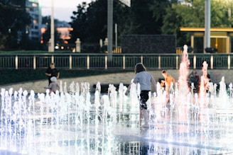 Children play around a lit fountain in an open public area during the evening. The water jets are illuminated with various colors, creating a vibrant and lively scene. In the background, there are people seated and relaxing, trees, and the surrounding cityscape with urban structures.
