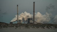 A large industrial facility with two tall smoke stacks releasing dense clouds of smoke into the overcast sky. The foreground includes grassy dunes that contrast with the industrial structures.