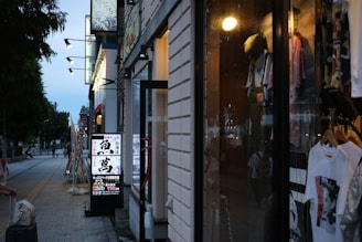 A city street scene at dusk with storefronts lit up. One shop window displays various clothing items on hangers. A brightly lit sign with Japanese text is visible, along with lamp posts lining the street. Trees and pedestrians walking with luggage add to the lively urban atmosphere.