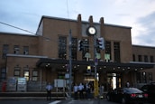 A brick building with large windows and a prominent clock above the entrance. Several people are waiting near the entrance, and a black car is parked nearby. Traffic lights and street signs are visible, and vending machines are placed to the side of the building.
