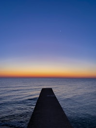 A jetty under construction extending into calm ocean waters at sunset.