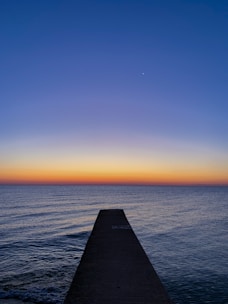 A jetty under construction extending into calm ocean waters at sunset.