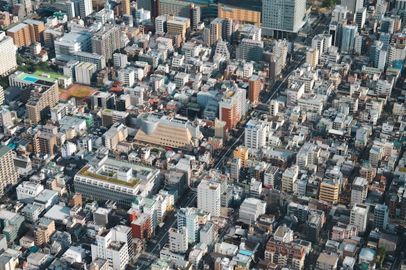 Aerial view of a densely populated urban area featuring numerous buildings of varying heights and architectural styles. The grid-like street layout separates residential and commercial structures. Green rooftops are visible on some buildings, alongside patches of green spaces such as sports fields.