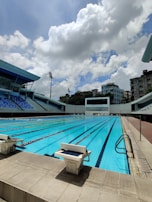 An outdoor swimming pool with clear blue water is surrounded by stadium seating. The sky is partly cloudy, creating a mix of sun and shade over the pool. Diving platforms are visible at the sides, and tall lighting fixtures rise above the seating area.