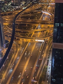An aerial view of a complex highway interchange with multiple lanes and overpasses, illuminated by street lights. Cars are visible on the roads, suggesting evening or nighttime traffic. Buildings and city lights are in the background, adding to the urban atmosphere.