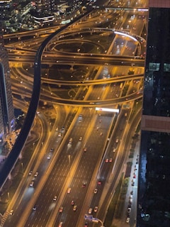 An aerial view of a complex highway interchange with multiple lanes and overpasses, illuminated by street lights. Cars are visible on the roads, suggesting evening or nighttime traffic. Buildings and city lights are in the background, adding to the urban atmosphere.
