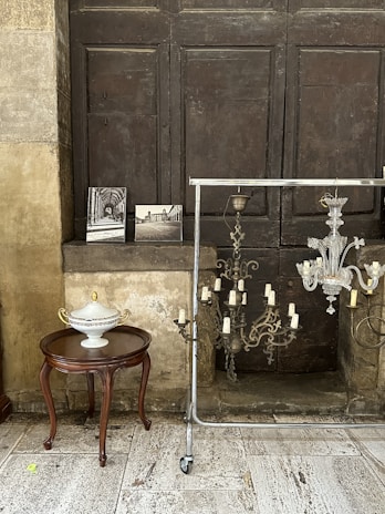 A vintage setting features a wooden table with a decorative ceramic urn placed on top. Next to it, several black and white photographs rest against a dark, wooden panel backdrop. A metal rod with wheels holds a chandelier with unlit candle holders. The flooring consists of light-colored stone tiles, and the entire arrangement exudes an old-world charm.