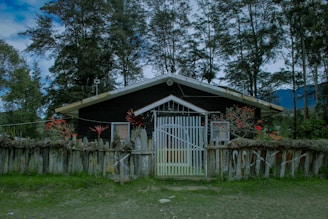 A wooden house surrounded by tall trees and a wooden fence. The house has a gabled roof, and the fence is decorated with red flowers and foliage. The sky appears overcast, and the area is lush with greenery.