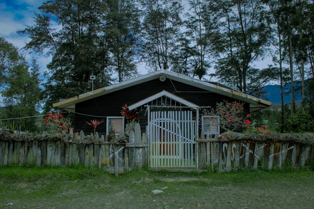 A wooden house surrounded by tall trees and a wooden fence. The house has a gabled roof, and the fence is decorated with red flowers and foliage. The sky appears overcast, and the area is lush with greenery.