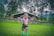 A child stands on a lush green lawn in front of a wooden house surrounded by tall trees. The child is wearing a colorful shirt, dark shorts, and bright green boots. The house has a rustic appearance with a fenced garden area.