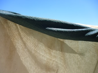 A close-up view of a partially transparent fabric hanging under a blue sky. The fabric, with a green edge, creates a shadow pattern as sunlight passes through its woven texture.