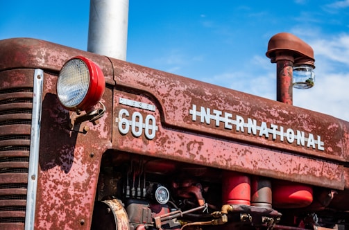 A vintage red tractor with the brand 'International' and model '600 Diesel' displayed in large letters. The tractor's surface shows signs of rust and wear, indicating age and use. The front light and various mechanical parts are visible. The sky is clear with a deep blue color, enhancing the rustic feel of the scene.