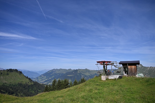 A scenic view of a mountainous landscape with lush green hills and a clear blue sky. There is a small wooden structure next to a ski lift or cable car station on the right side of the image. In the background, layers of mountains stretch into the distance.